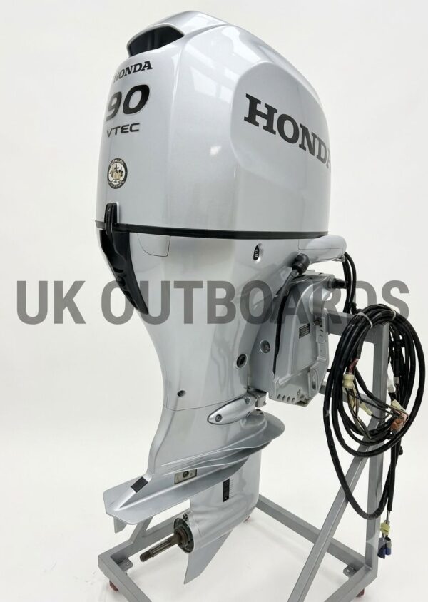 Pristine Honda BF90 EFI - 2021 90HP Four-Stroke Remote Outboard Motor Three-quarter rear view of a pristine honda bf90 efi outboard motor resting on a workshop rigging stand, showcasing its factory-fresh metallic silver paint, sleek dark gray decals, and unpolished stainless steel propeller.